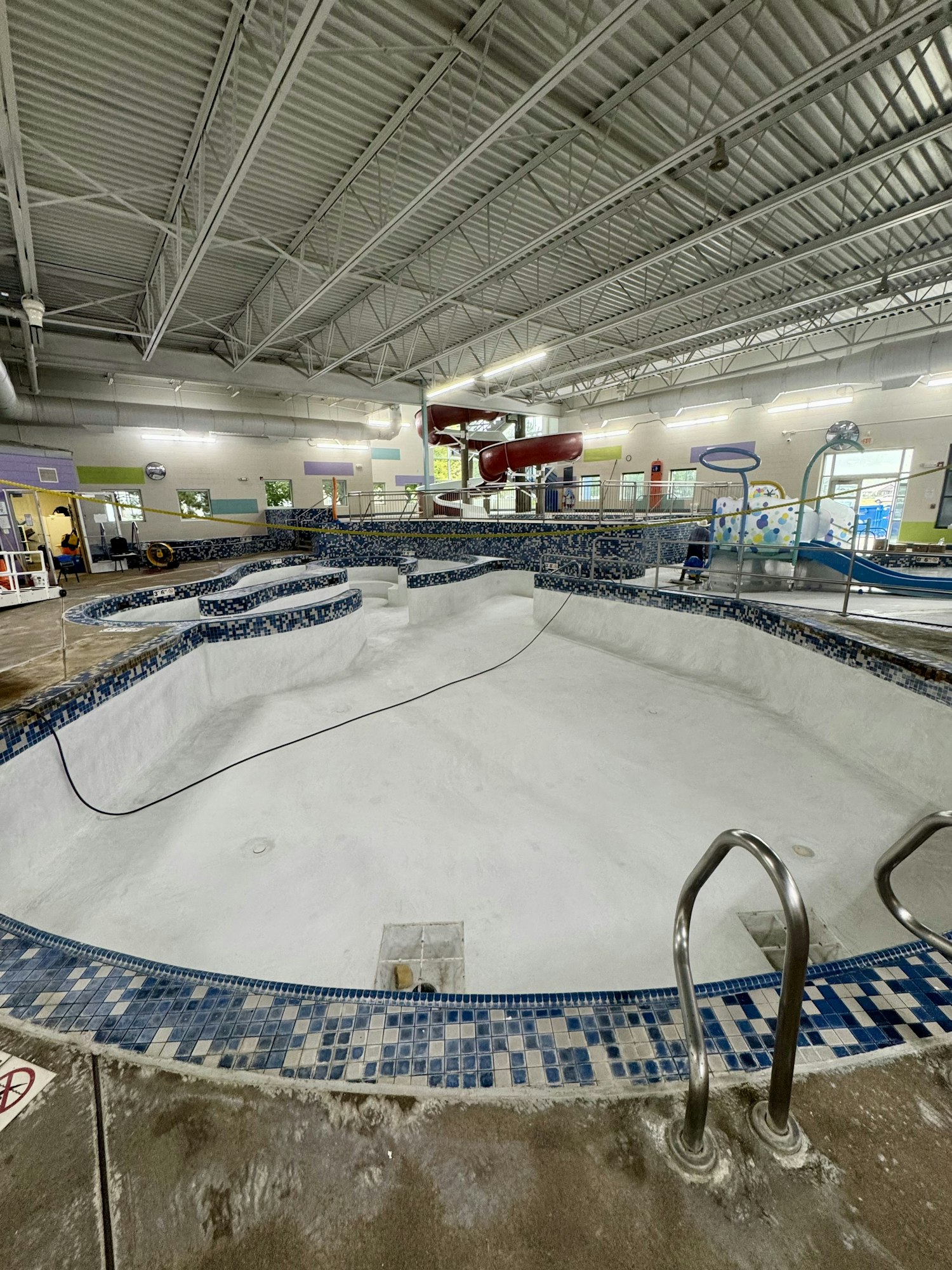 An empty indoor pool with a waterslide, featuring blue tile edges and a high ceiling with exposed metal beams.