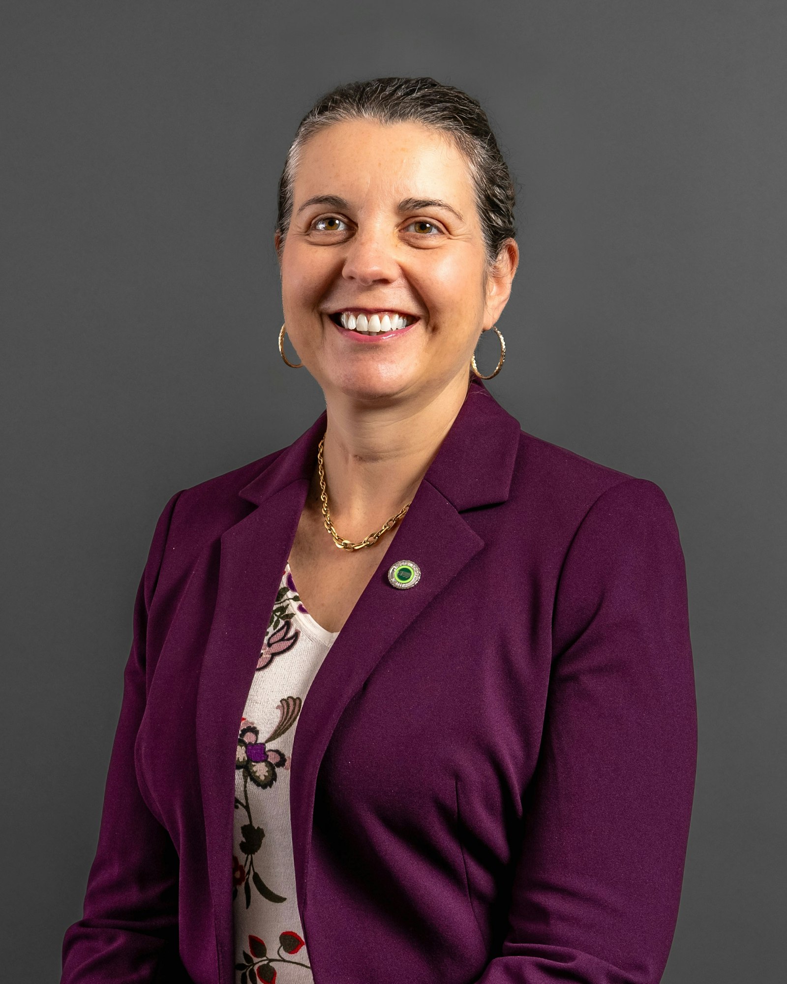 A person smiling, wearing a purple blazer and floral shirt, against a gray background.