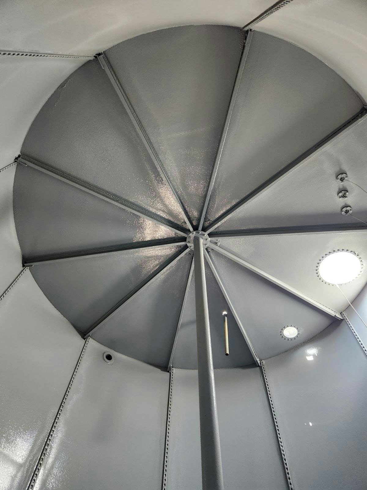 Interior of a metallic structure, possibly a silo or tank, with rivet patterns, light fixtures, and a vertical pipe.