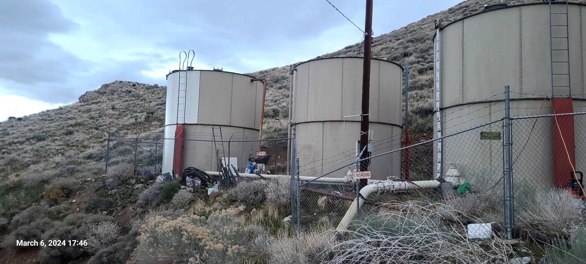 Industrial storage tanks behind a chain-link fence with "No Trespassing" signs, amidst a natural, hilly terrain.