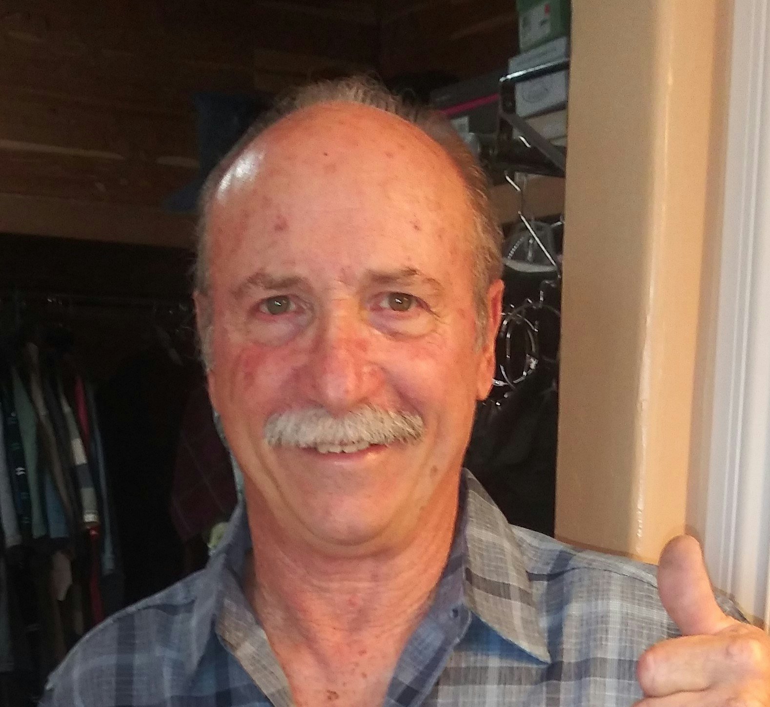 A smiling man with a mustache gives a thumbs-up, standing in front of a closet with clothes in the background.
