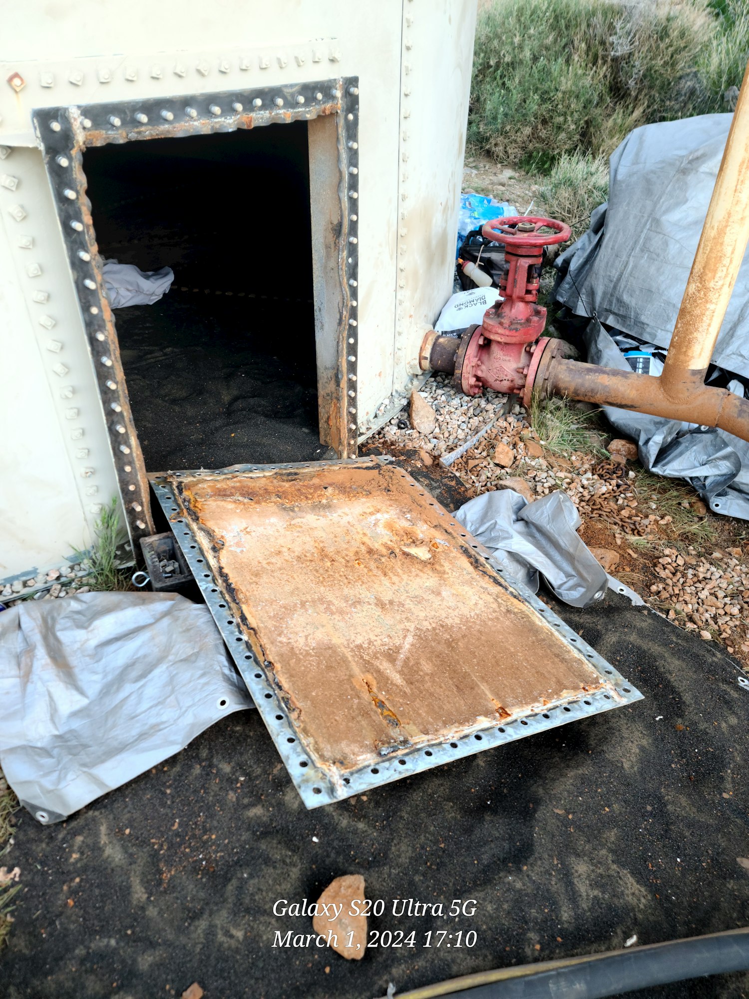 Image of an access door lying on the ground in front of an empty water storage tank