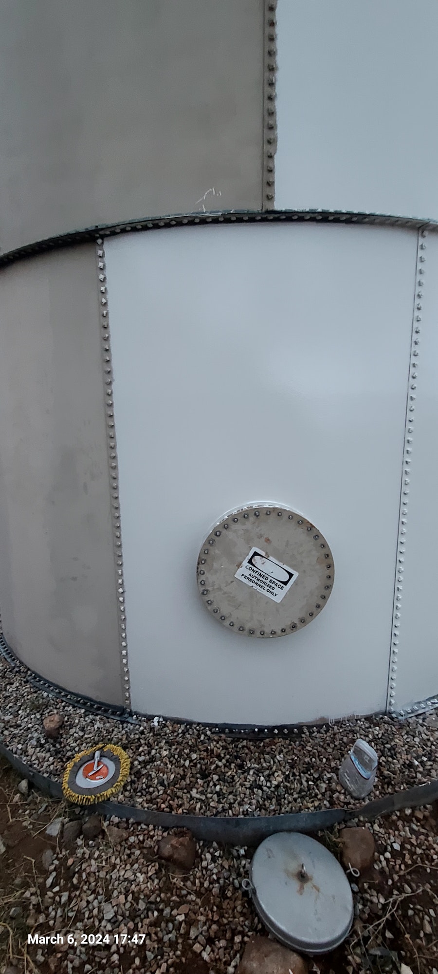 An industrial storage tank with riveted seams, inspection cover, and gravel surround, viewed from an angle.