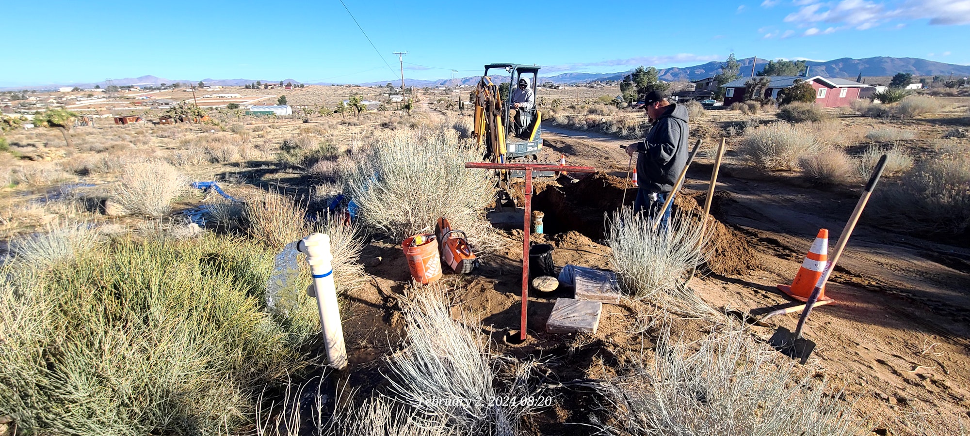 A construction site with workers, an excavator, a dug trench, safety cones, tools, and rural surroundings.