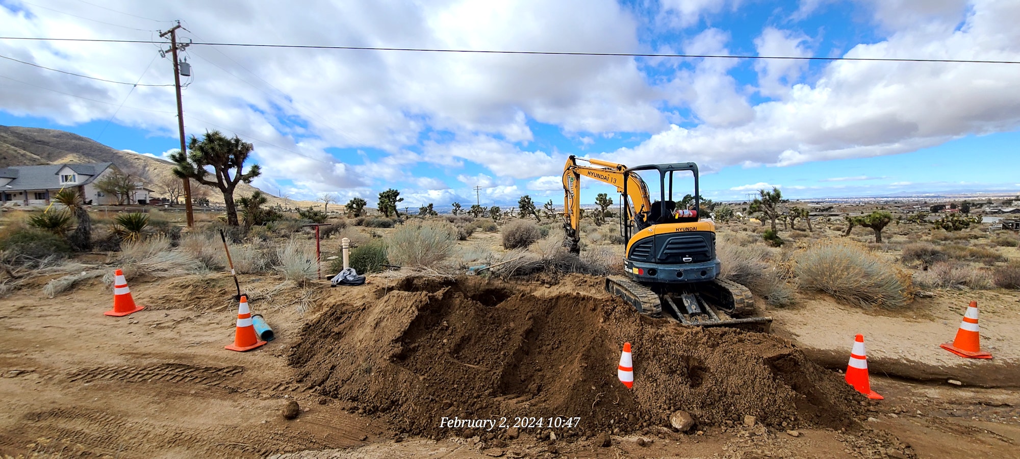 A mini excavator on a construction site with traffic cones, desert vegetation, and a cloudy sky.