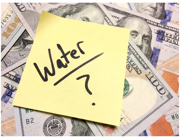 A sticky note saying "Water?" is placed on a pile of U.S. hundred-dollar bills, suggesting a question about water's value or cost.