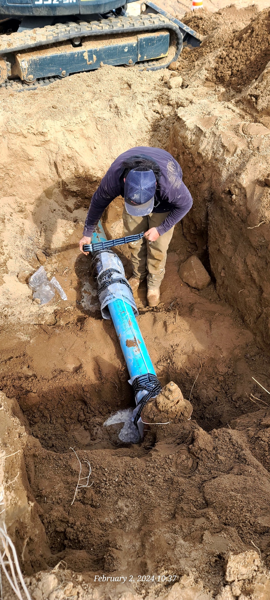 A person in a cap is working on blue underground pipes in a dirt trench, with excavator tracks visible above.