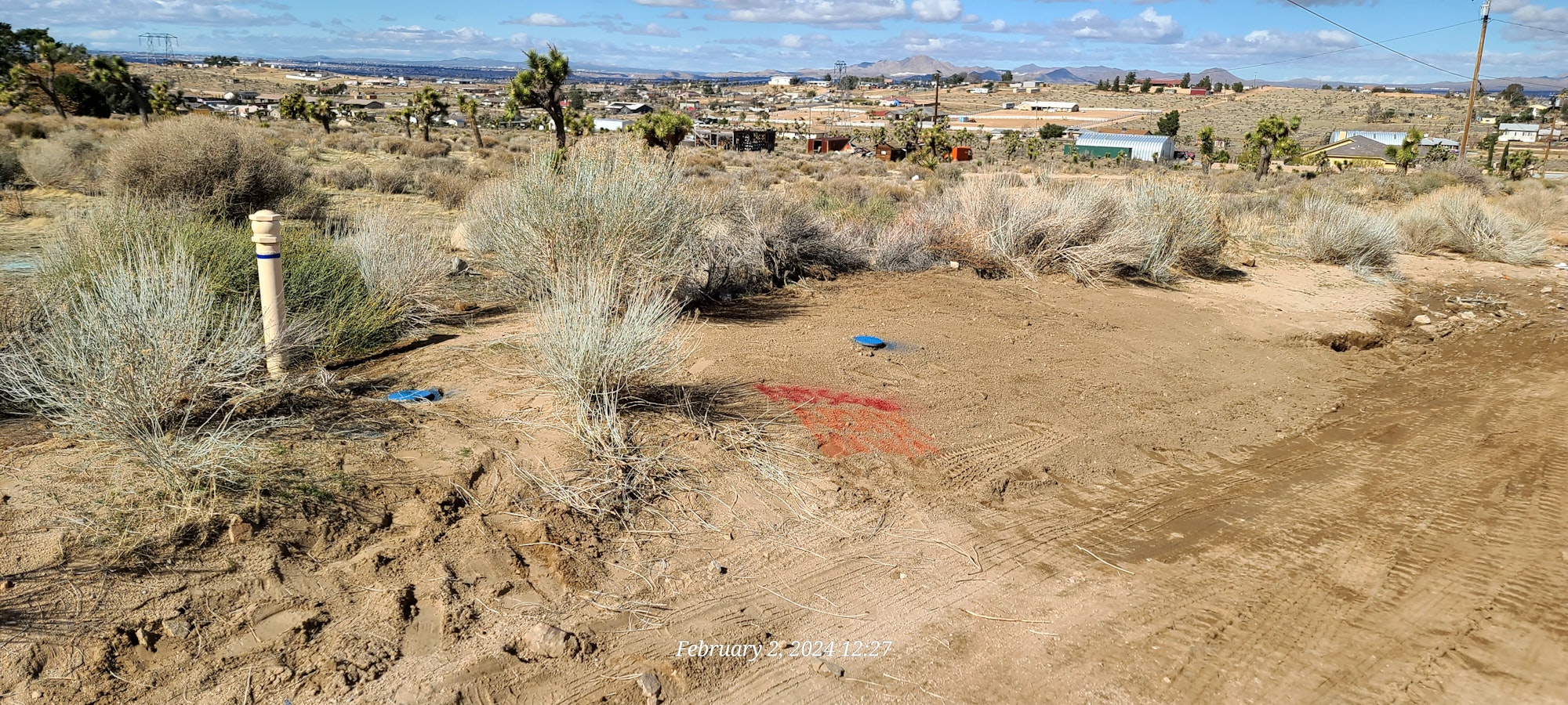 Desert terrain with sparse vegetation, a marked utility pole, tire tracks, under a cloudy sky.