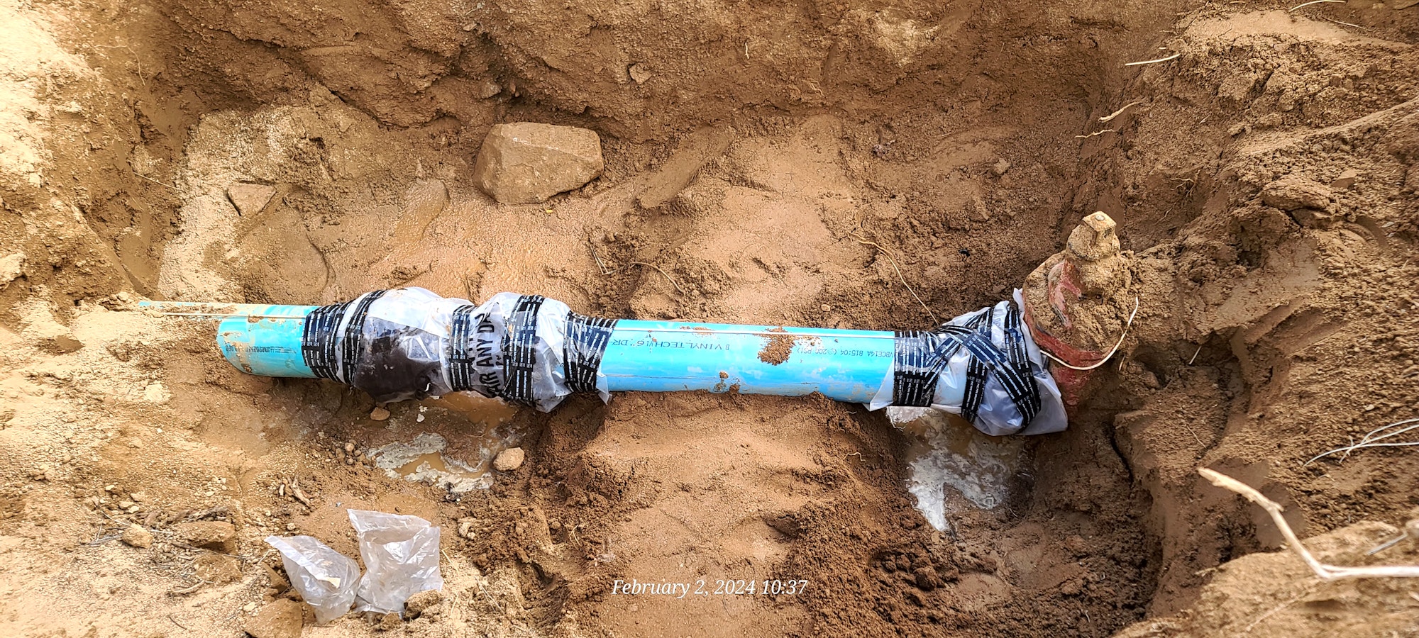 A repaired blue water pipe in a trench, wrapped with tape and plastic.