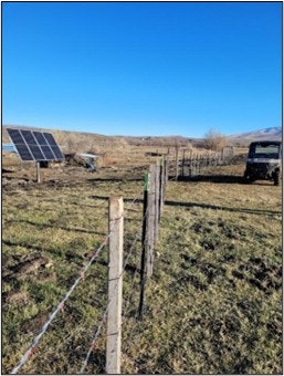 The image shows a rural landscape with a solar panel, a fence, grassy land, and an off-road vehicle under a clear blue sky.
