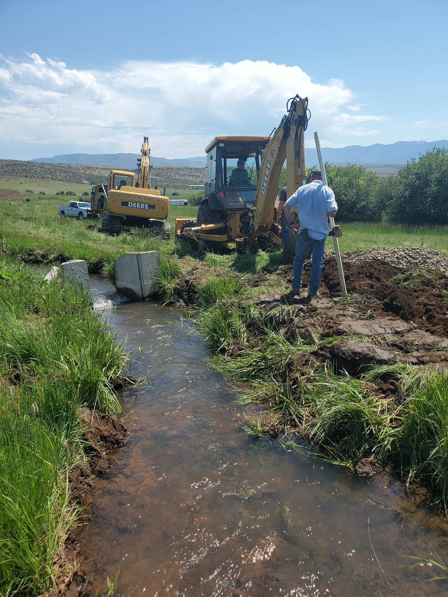 Construction equipment working by a stream, with workers managing the site surrounded by grass and hills under a clear sky.