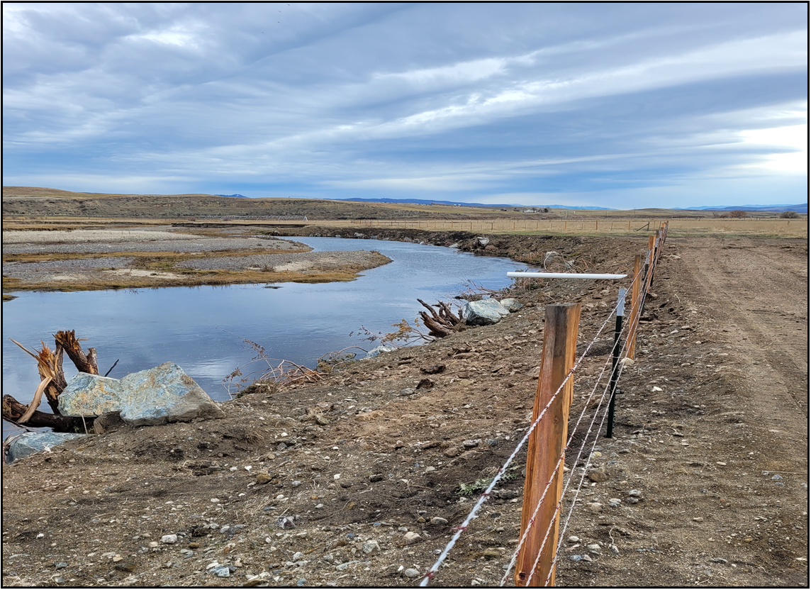 A meandering river next to a dirt road with a wire fence under a blue sky with wispy clouds. Rural landscape with no people.