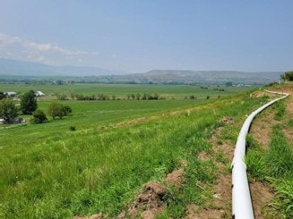 The image shows a grassy landscape with rolling hills, fields, and a white pipe running through the foreground.