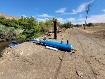 A blue water pump is located near a riverbank, surrounded by dry land and vegetation, with power lines in the background.
