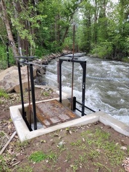 A metal structure with a flat surface near a flowing river, likely a water control or observation station in a natural setting.