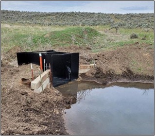 A black structure near a pond, surrounded by earth and grass, likely a water management or drainage system.