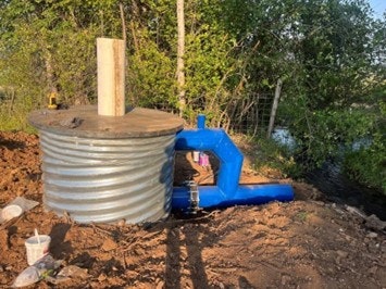 The image shows a large metal tank with a blue pipe attached, situated near a body of water and surrounded by greenery.