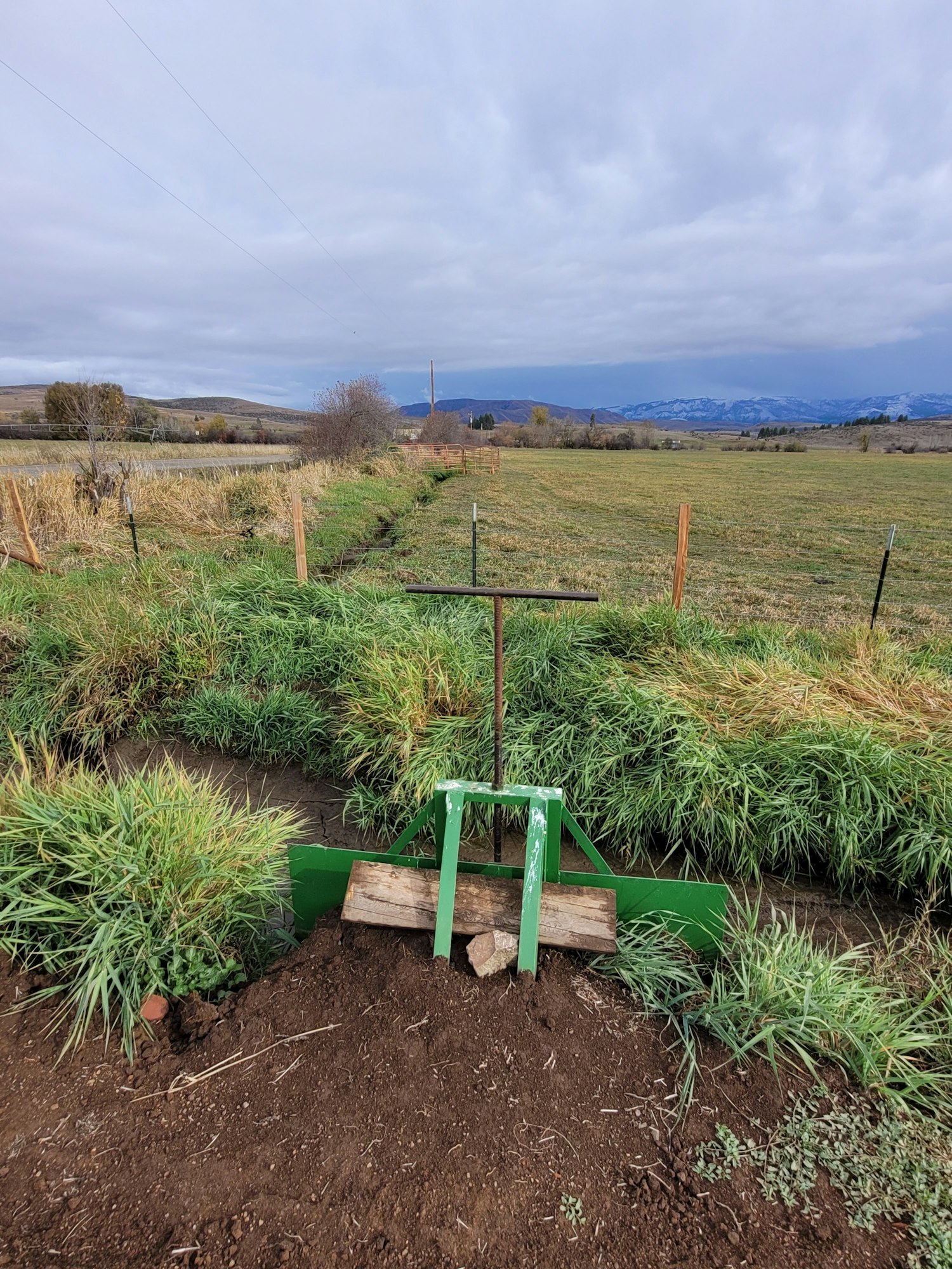 Rural landscape with green wheelbarrow, wooden planks, cultivated soil, grassy field, and cloudy sky. (Image is rotated)
