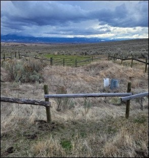 A wide landscape with grasslands, a few wooden fences, and a cloudy sky. A small structure is visible in the grass.