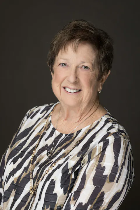 A smiling elderly woman wearing a patterned blouse and jewelry poses against a dark background.