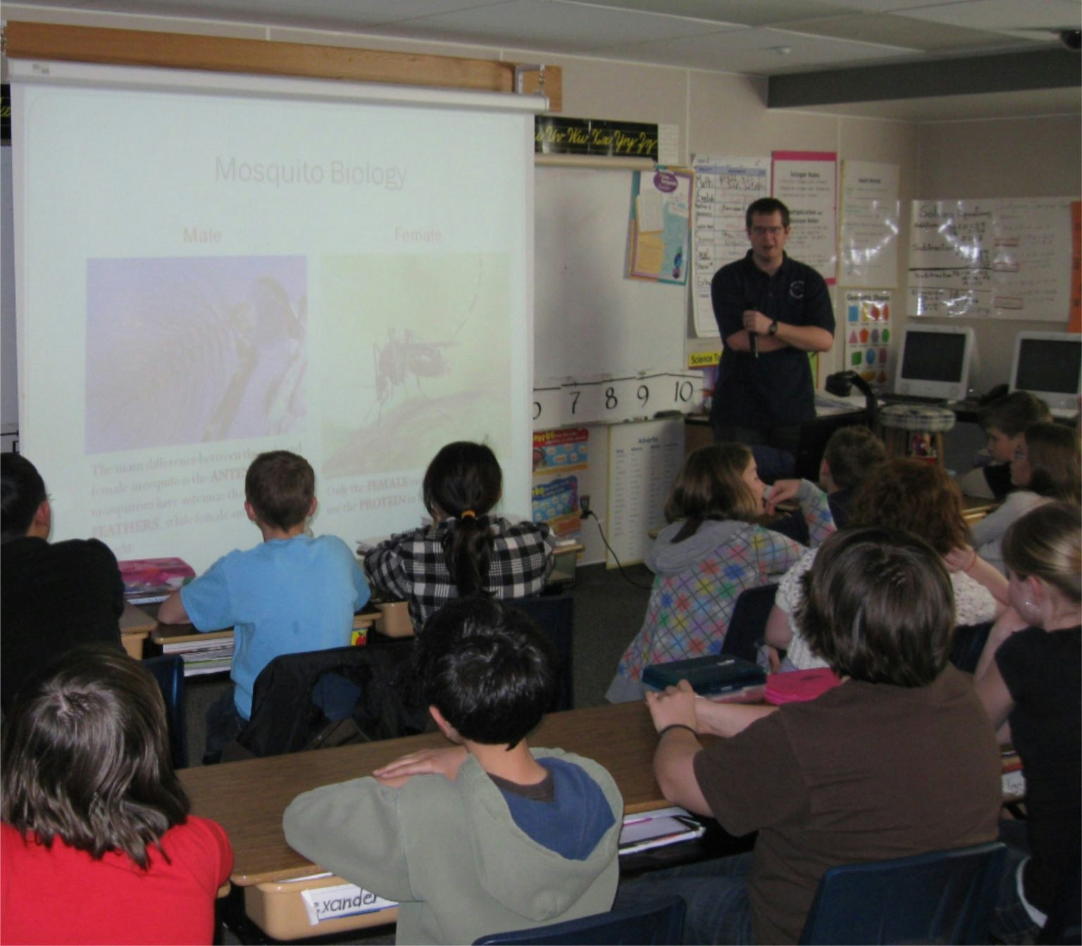 A classroom with students focused on a "Mosquito Biology" presentation, and a teacher standing on the side.