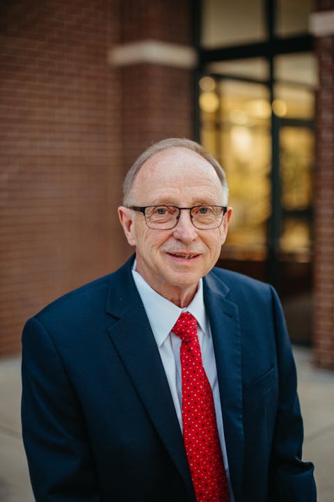 Elderly man in glasses, wearing a suit and red tie, standing outside a brick building.