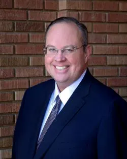 A smiling man in a suit stands against a brick wall, exuding a professional demeanor.