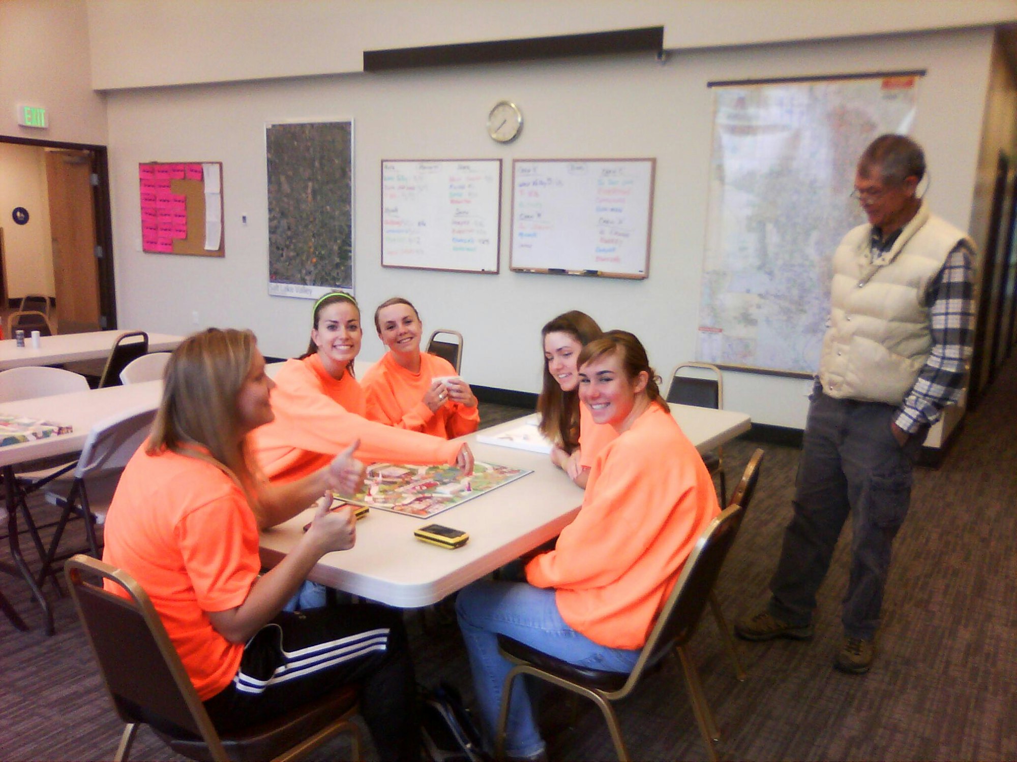 Five people in a room, four in orange shirts are playing a board game, one man is standing beside them, observing.
