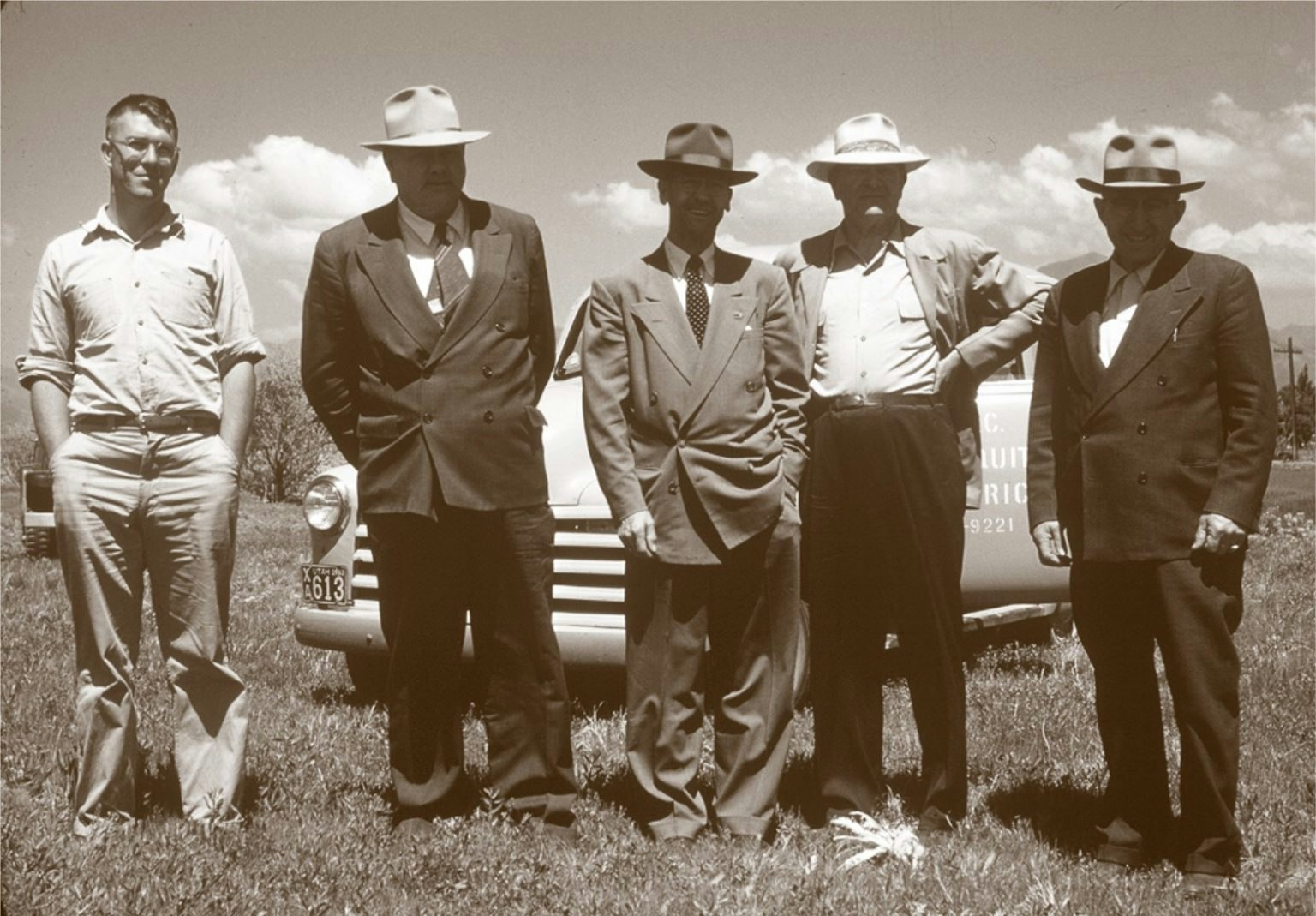 Five men standing in front of a vintage car, four wearing hats and suits, one in casual attire.