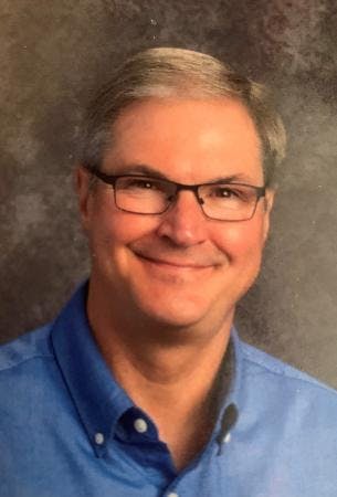 A smiling person wearing glasses and a blue shirt, with a gray background.