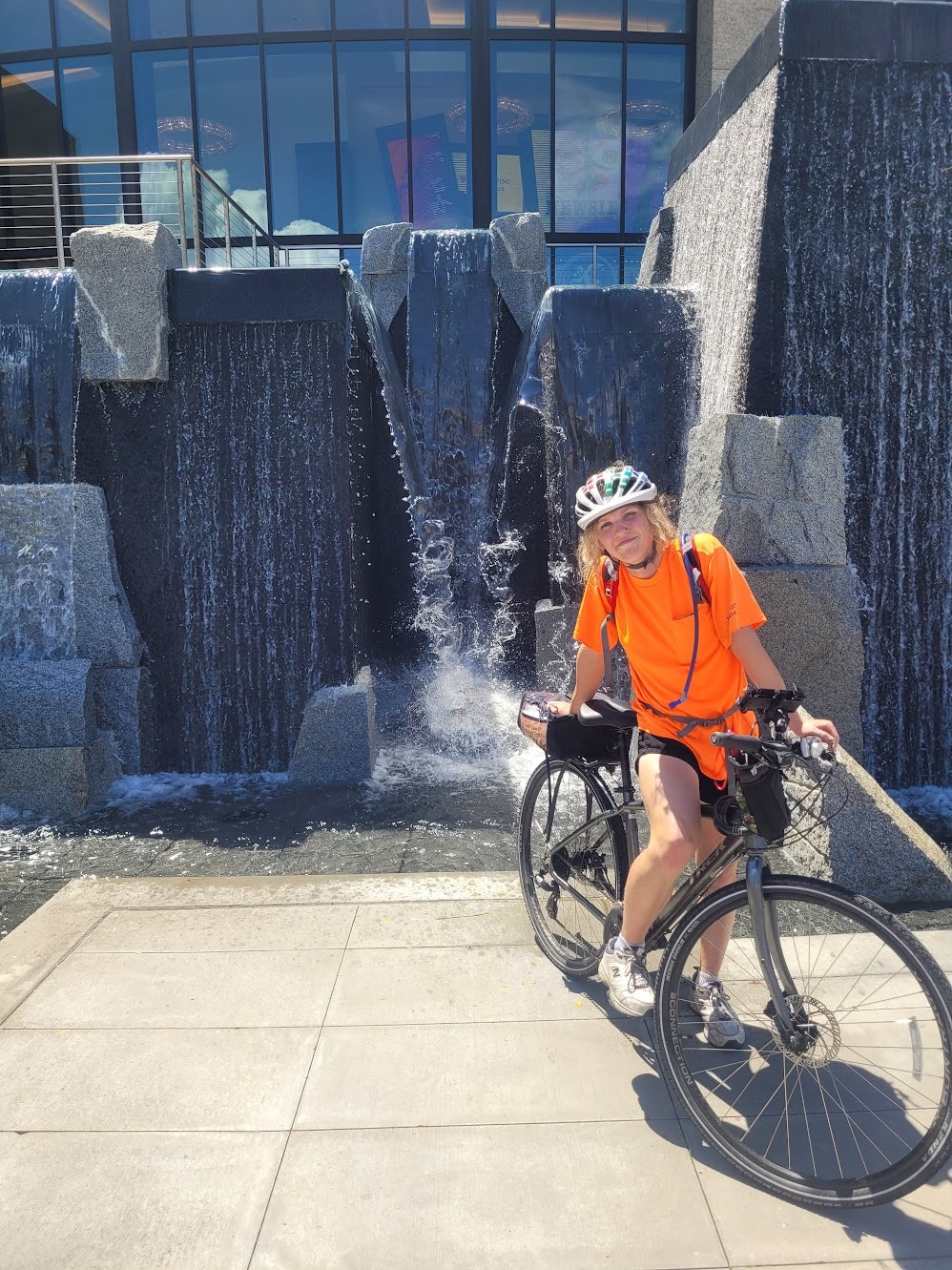 A person in an orange shirt and helmet poses with a bike in front of a modern fountain.