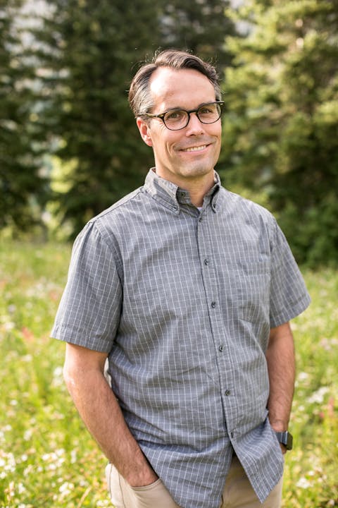 A person wearing glasses and a checkered shirt, standing outdoors with trees in the background.