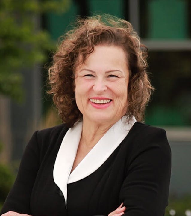 A person with curly hair, smiling, wearing a black outfit with a white collar, standing outdoors.