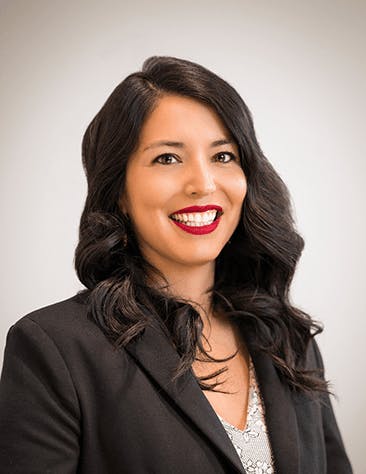 A person with long dark hair and a black blazer smiles at the camera against a plain background.
