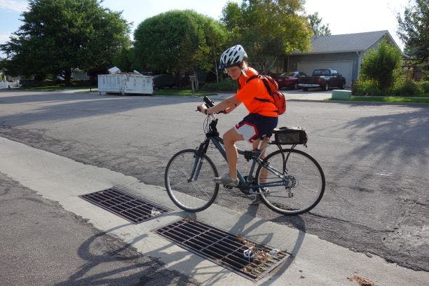A person in a helmet and orange shirt is riding a bicycle over street drains on a sunny day.