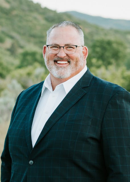 A smiling man in a suit and glasses stands outdoors with a scenic, green background.