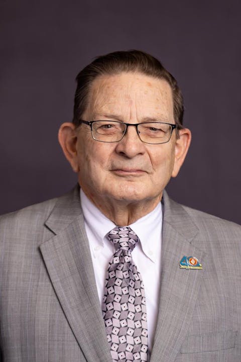 An older man wearing a gray suit, patterned tie, and glasses, with a lapel pin, set against a plain dark background.