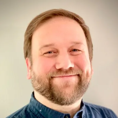 The image features a smiling person with a beard, wearing a blue shirt, against a plain background.