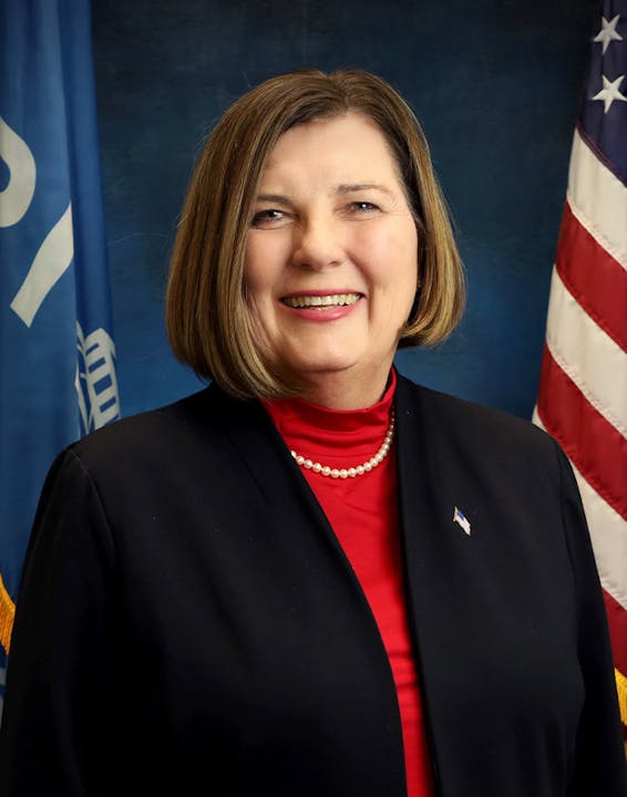 A person smiling, wearing a red top and black jacket, with U.S. and blue flags in the background.