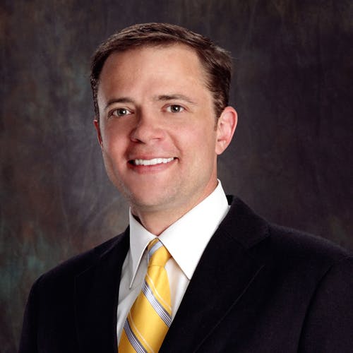 A man in a suit and yellow tie, smiling, with a neutral background.