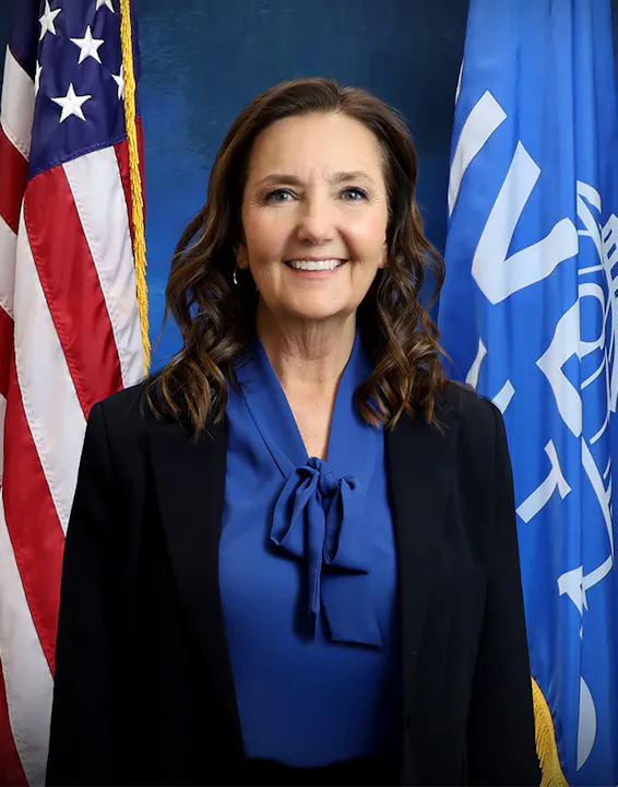 A woman in a blue blouse and black blazer smiles, standing in front of U.S. flags and a blue backdrop.