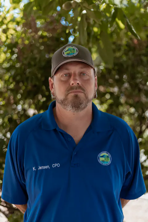 A person in a blue shirt and cap stands outdoors, with trees in the background.
