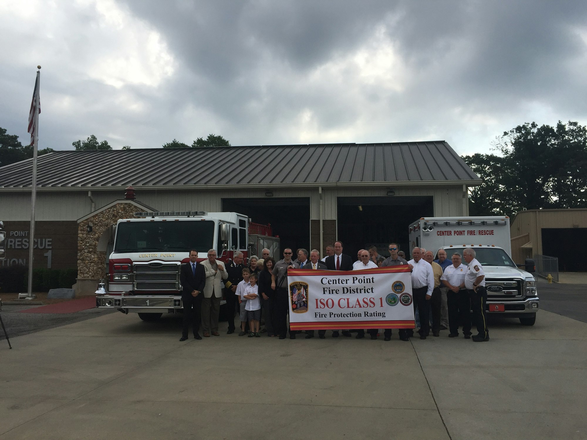 A group of people stand in front of a fire station, holding a banner for Center Point Fire District's ISO Class 1 Fire Protection Rating.