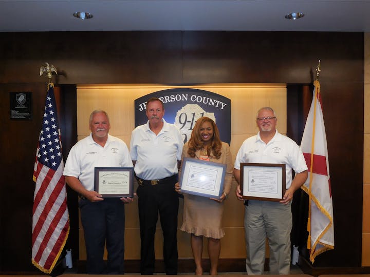 Four people holding certificates in front of a "Jefferson County 9-1-1" sign with American flags.