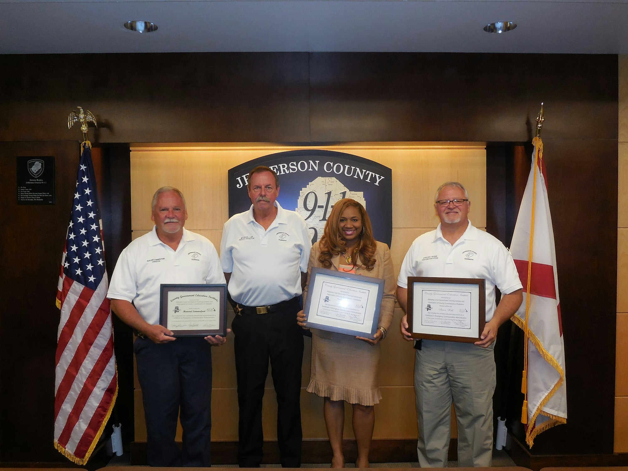 Four people holding certificates in front of a "Jefferson County 9-1-1" sign with American flags.