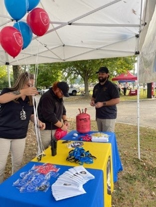 A table under a canopy with balloons, leaflets, keychains, two people setting up, and one person standing nearby.