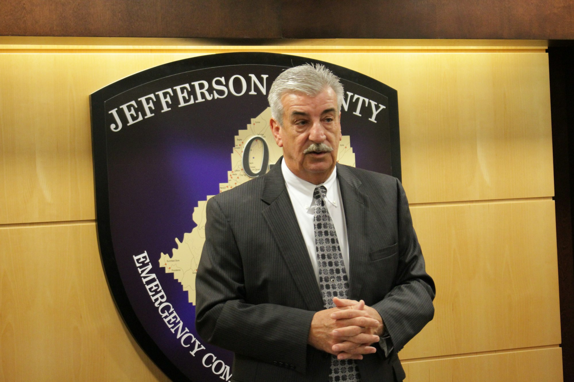 A man in a suit stands in front of a sign reading "Jefferson County Emergency".