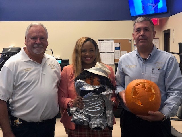 Three people pose indoors; one holds a pumpkin, another holds a decorated object. They appear to be in an office setting.