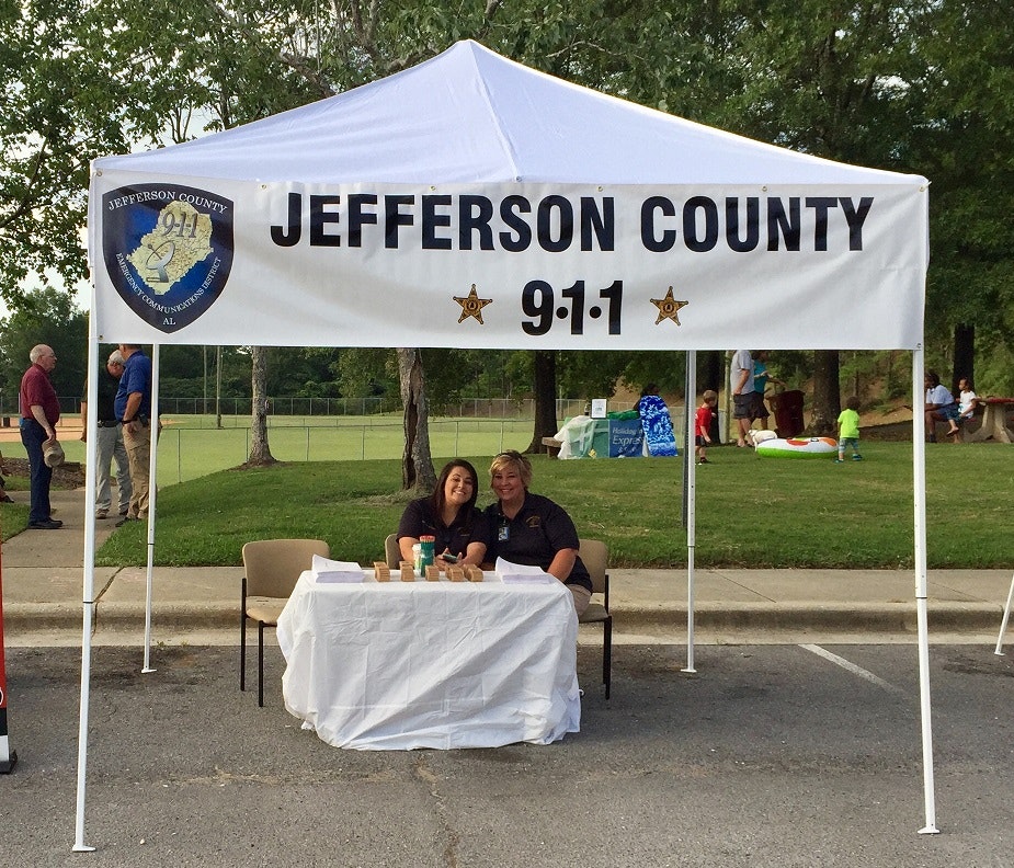 A booth under a tent with a "Jefferson County 9-1-1" sign and two people sitting at a table.