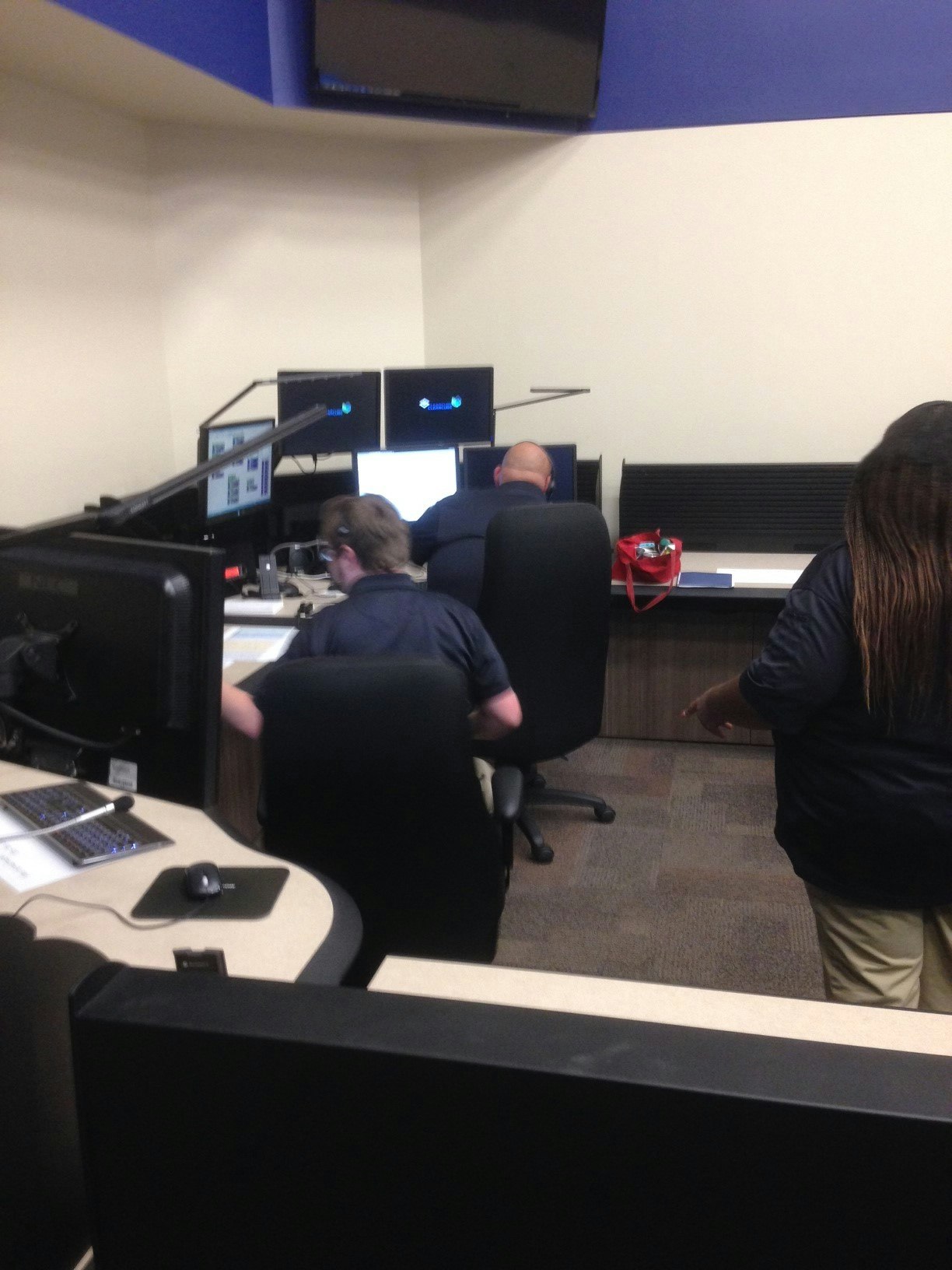 People in an office setting working at computer stations with multiple monitors and a red bag on the desk.